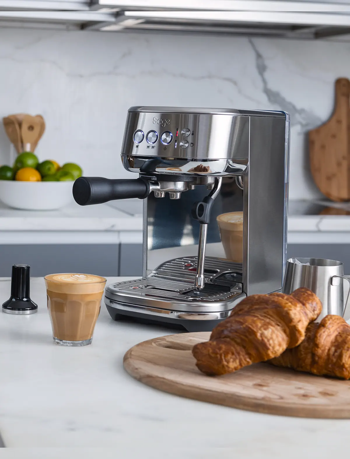 Cafetera Sage Bambino Inox en una cocina con croissants y un café cortado con crema perfecta en vaso de cristal.